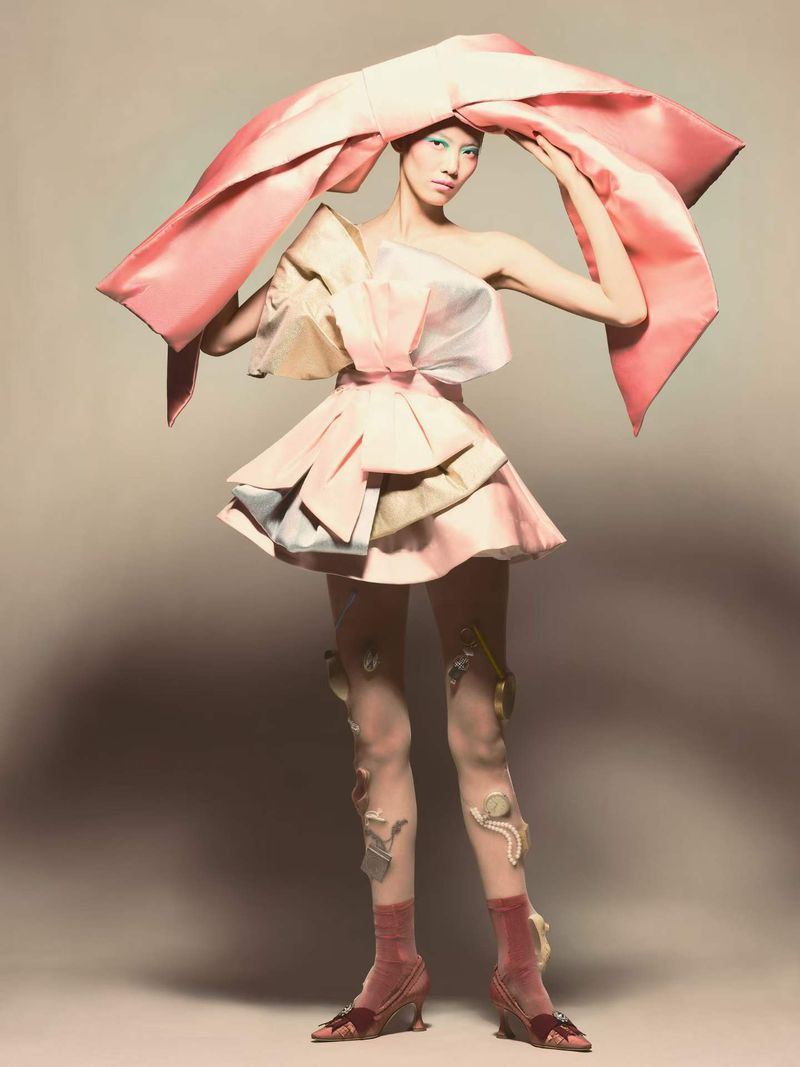 Model Hejia Li wearing an oversized pink bow headpiece and sculptural mini dress against a neutral backdrop.