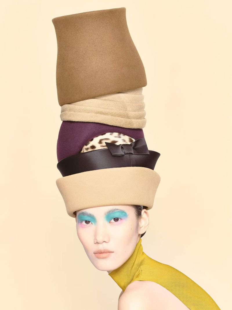 Model Hejia Li balances a tall sculptural stack of felt and leather hats against a pale background for W Magazine China.