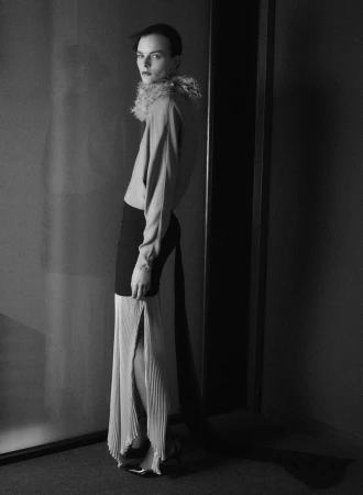 Model Aylah Peterson in a ruffled collar top and pleated slit skirt standing in profile in a monochrome studio setting.