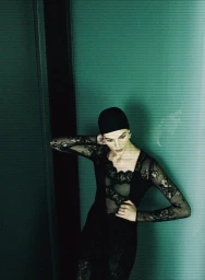 Model Aylah Peterson in a black lace dress and black cap posing against a green wall in an editorial fashion shoot.