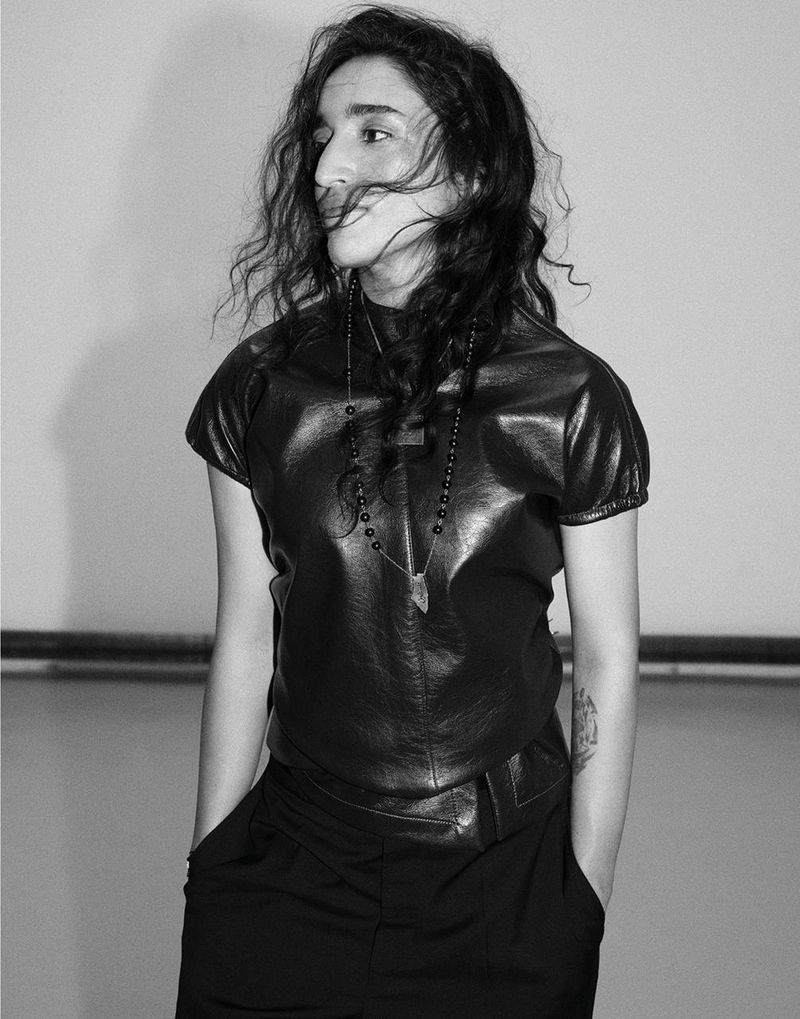 Black and white portrait of Sama' Abdulhadi wearing a short-sleeve leather top and beaded necklace with hands in pockets.