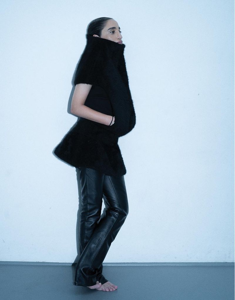 Palestinian DJ Sama' Abdulhadi posing barefoot in a sculptural black high-neck top and black leather pants against a pale backdrop.