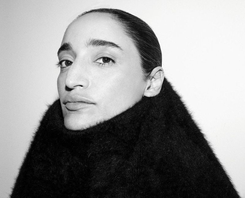 Black and white close-up portrait of DJ Sama' Abdulhadi looking at camera, wearing a thick textured black high-necked garment against a white background.