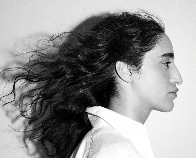 Black and white profile portrait of Sama' Abdulhadi with windswept dark hair wearing a white collared shirt against a light wall.