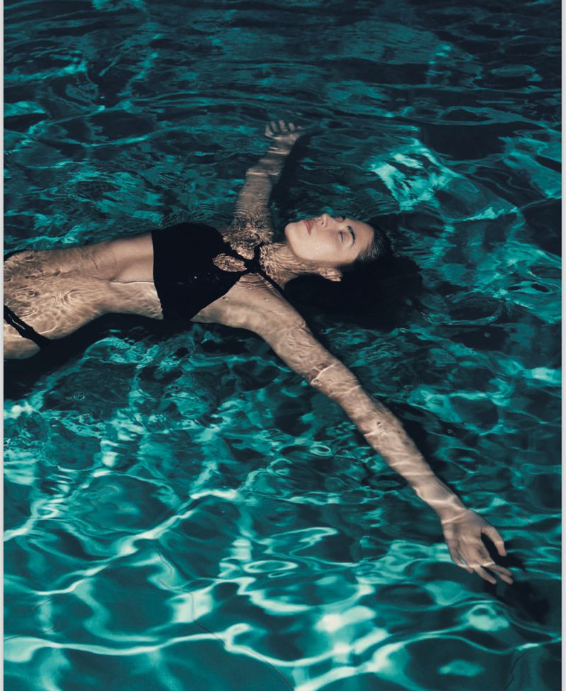 Woman in black bikini floats in a pool with teal water. Her eyes are closed, and her arms are outstretched. The pool water reflects light.