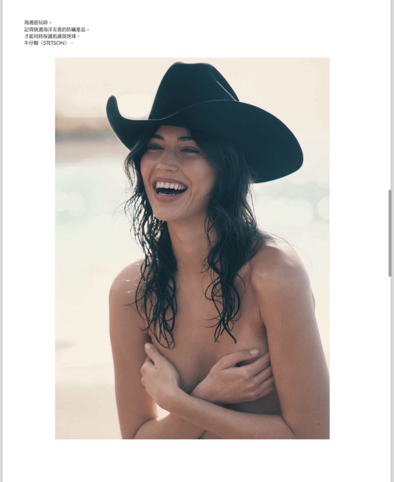 Smiling woman wears a black cowboy hat, covering her chest with hands, enjoying a sunny outdoor setting with sandy background.