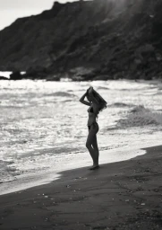 Model standing on a sandy beach by the ocean, holding her hair as waves break behind her under sunlight.