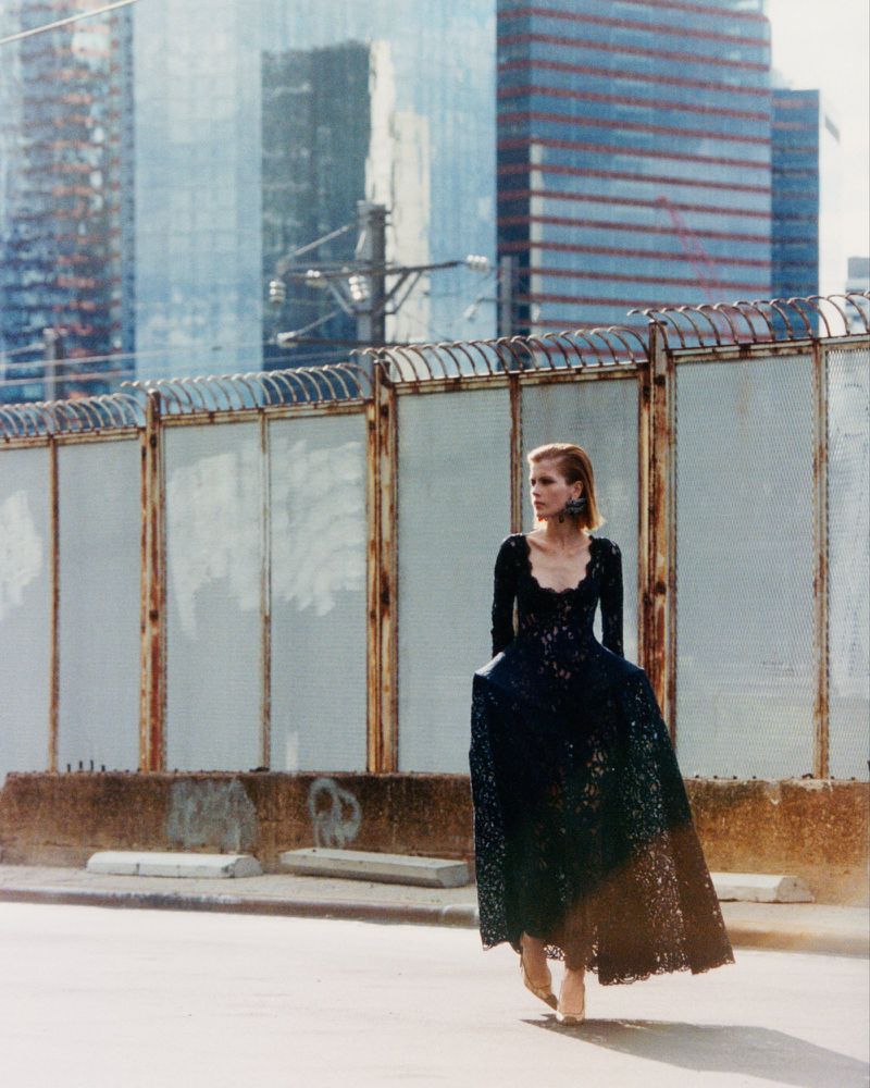 A woman wearing a floor-length black lace gown walking on a street in front of a barbed wire fence and skyscrapers.