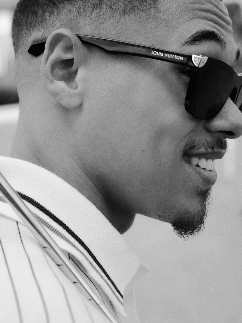 Close-up profile of actor Eliyha Altena smiling while wearing black Louis Vuitton sunglasses and a white collared pinstripe shirt.
