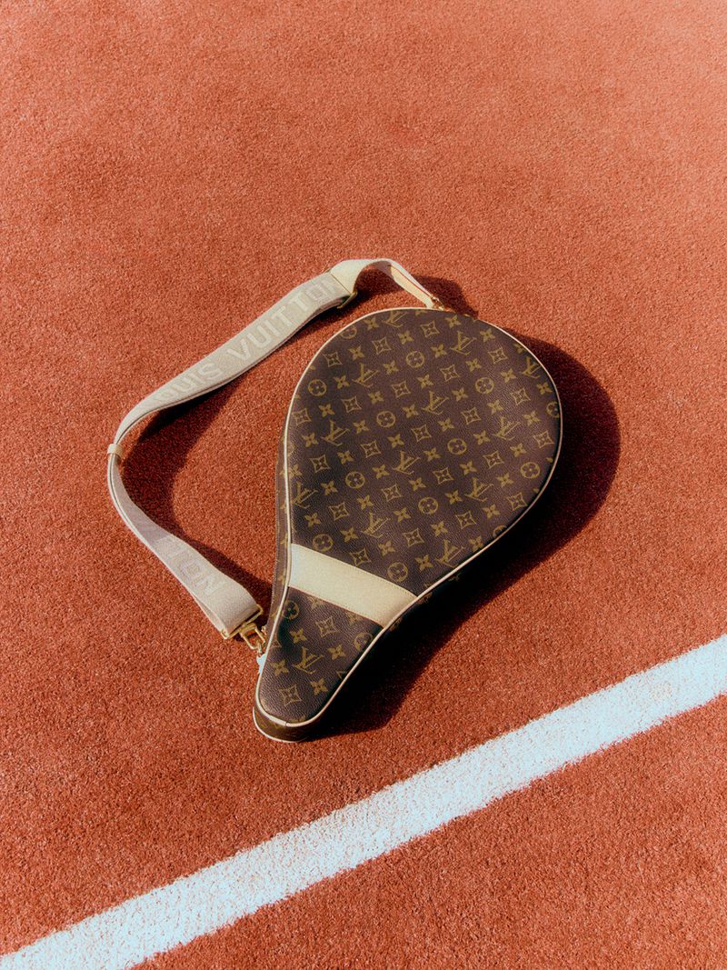 A brown monogram tennis racket cover with a cream shoulder strap resting on a red clay tennis court.