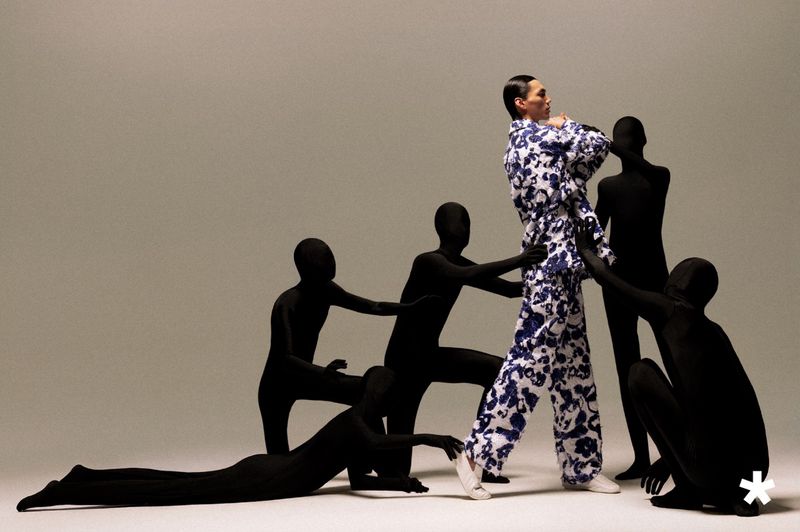 Model in a blue and white floral patterned suit walking while surrounded by figures in full-body black bodysuits in a studio.