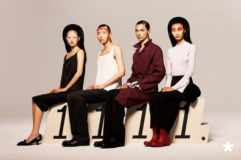 Four fashion models with avant-garde makeup and hairstyles seated on a white plinth marked 1111 against a seamless backdrop.