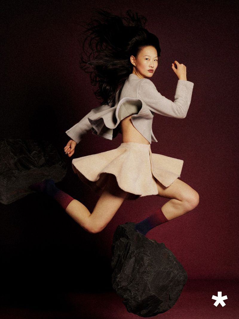 East Asian model in a sculptural flared mini skirt and cropped jacket leaping mid-air above dark floating rocks in a studio setting.