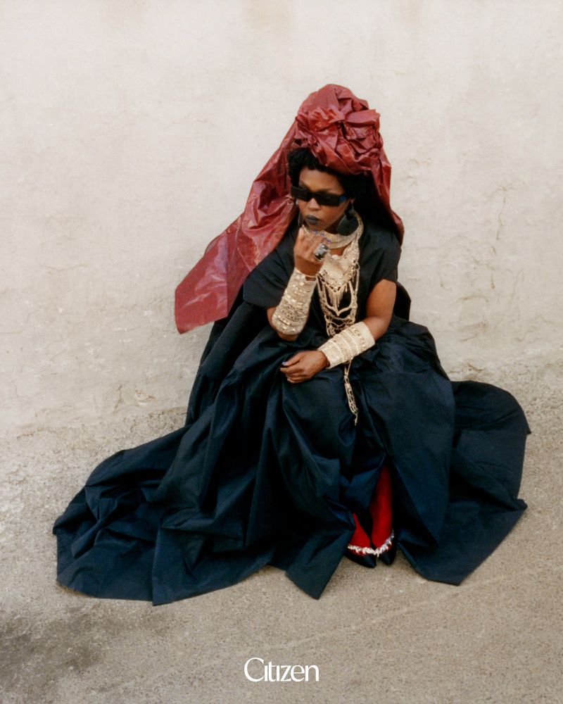 Lauryn Hill seated in a voluminous black gown, dramatic red headpiece, and sunglasses against a textured pale wall.