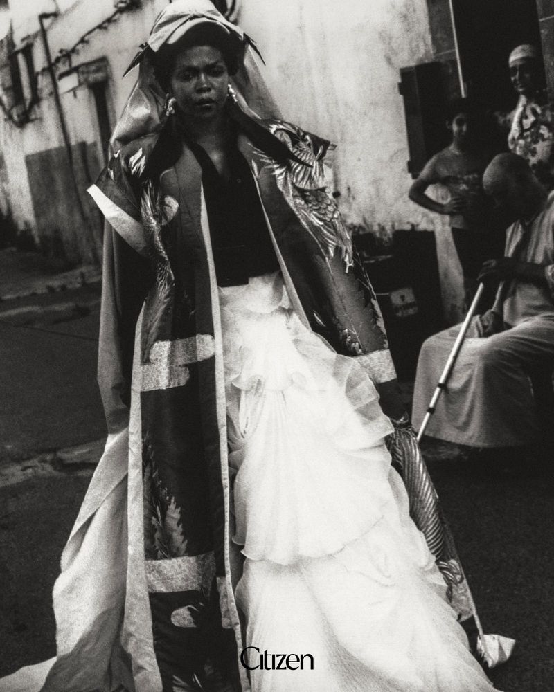 Lauryn Hill wearing a voluminous tiered white gown and patterned coat on a street in a black and white editorial portrait.