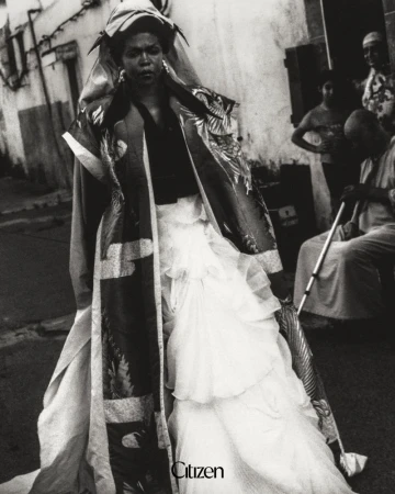 Lauryn Hill wearing a voluminous tiered white gown and patterned coat on a street in a black and white editorial portrait.