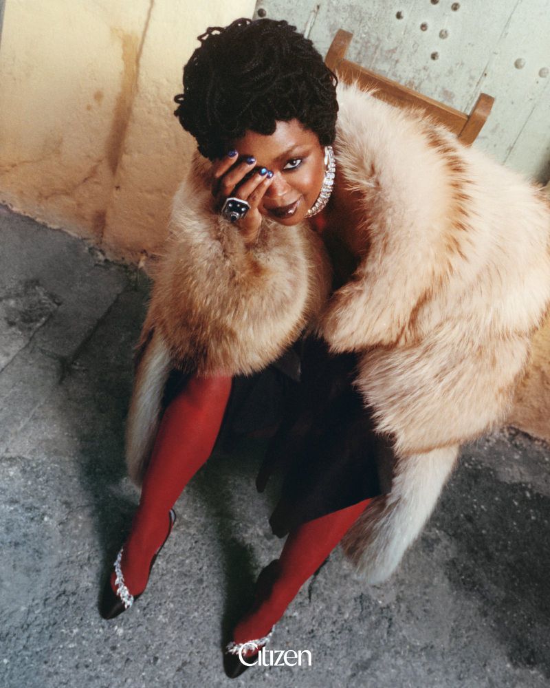 Ms. Lauryn Hill seated in a voluminous fur coat, red tights, and embellished heels for Citizen Magazine.