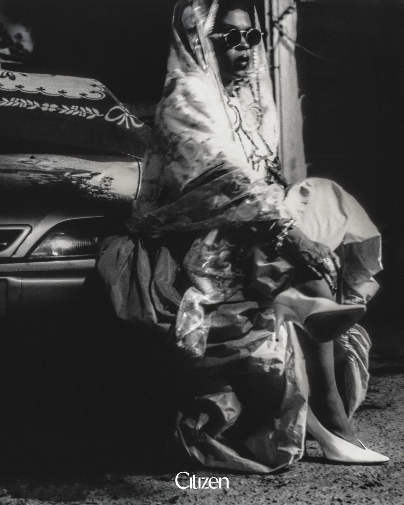 Lauryn Hill wearing sunglasses, a draped patterned shawl, and white heels, seated against a car in a black and white portrait.