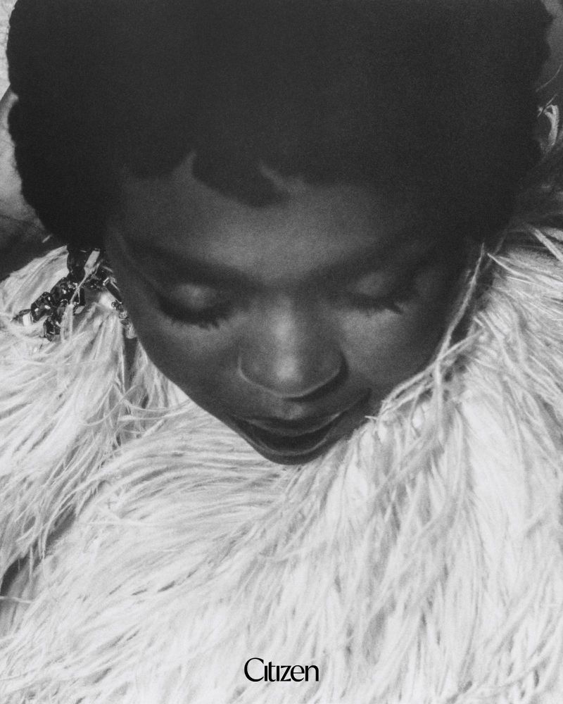 Black and white portrait of Ms. Lauryn Hill looking down, wearing a voluminous white feathered collar with the Citizen logo below.