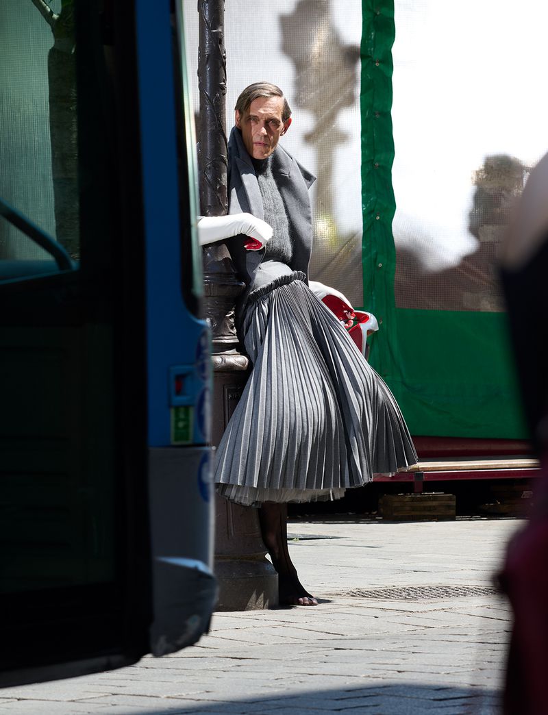 Model in an exaggerated grey pleated skirt and tailored grey jacket leaning against a street lamppost in bright daylight.
