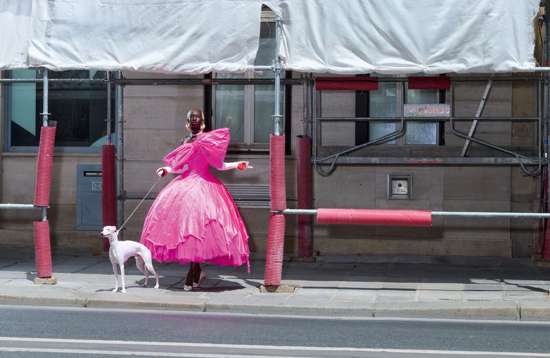 Model in an oversized bright pink ruffled gown walking a white greyhound on a city sidewalk beside scaffolding.