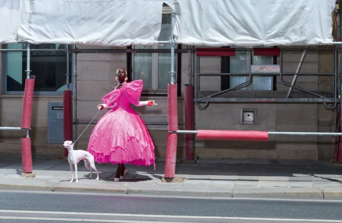 Model in an oversized bright pink ruffled gown walking a white greyhound on a city sidewalk beside scaffolding.