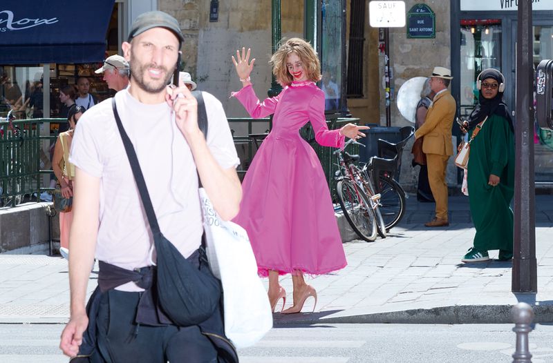 Model posing in a bright pink structured dress and sculptural high heels on a sunny Paris street with pedestrians.