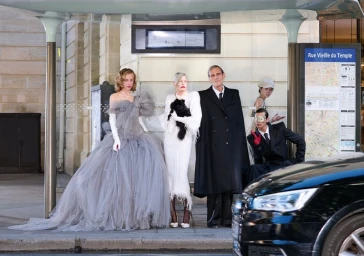 Fashion models in avant-garde couture gowns and tailored black coats posing together inside a Parisian bus stop.