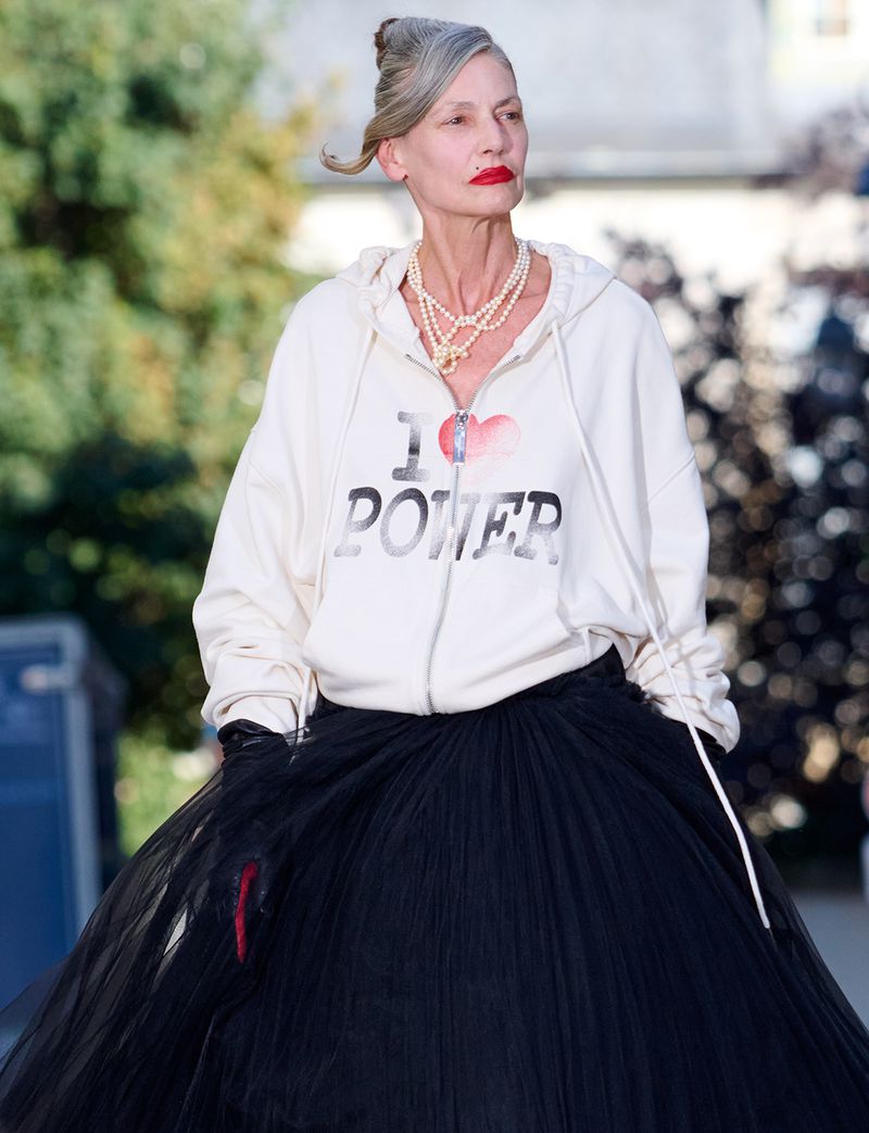 Mature model wearing an I Heart Power white zip hoodie, pearl necklace, and voluminous black tulle skirt in outdoor daylight.
