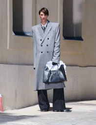 A male model wearing a structured grey double-breasted overcoat and wide black trousers holding a black leather tote on a sunlit street.