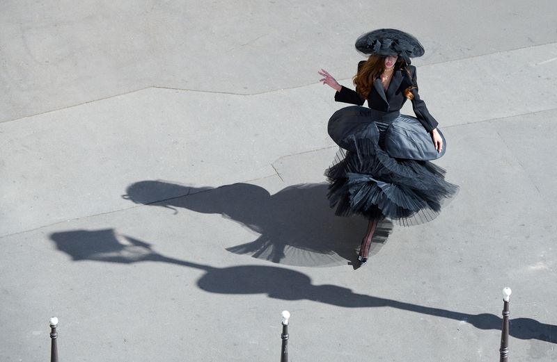 High-angle view of a woman wearing an avant-garde black tiered tulle dress and wide-brim hat walking across sunlit pavement.