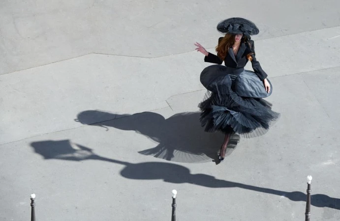 High-angle view of a woman wearing an avant-garde black tiered tulle dress and wide-brim hat walking across sunlit pavement.