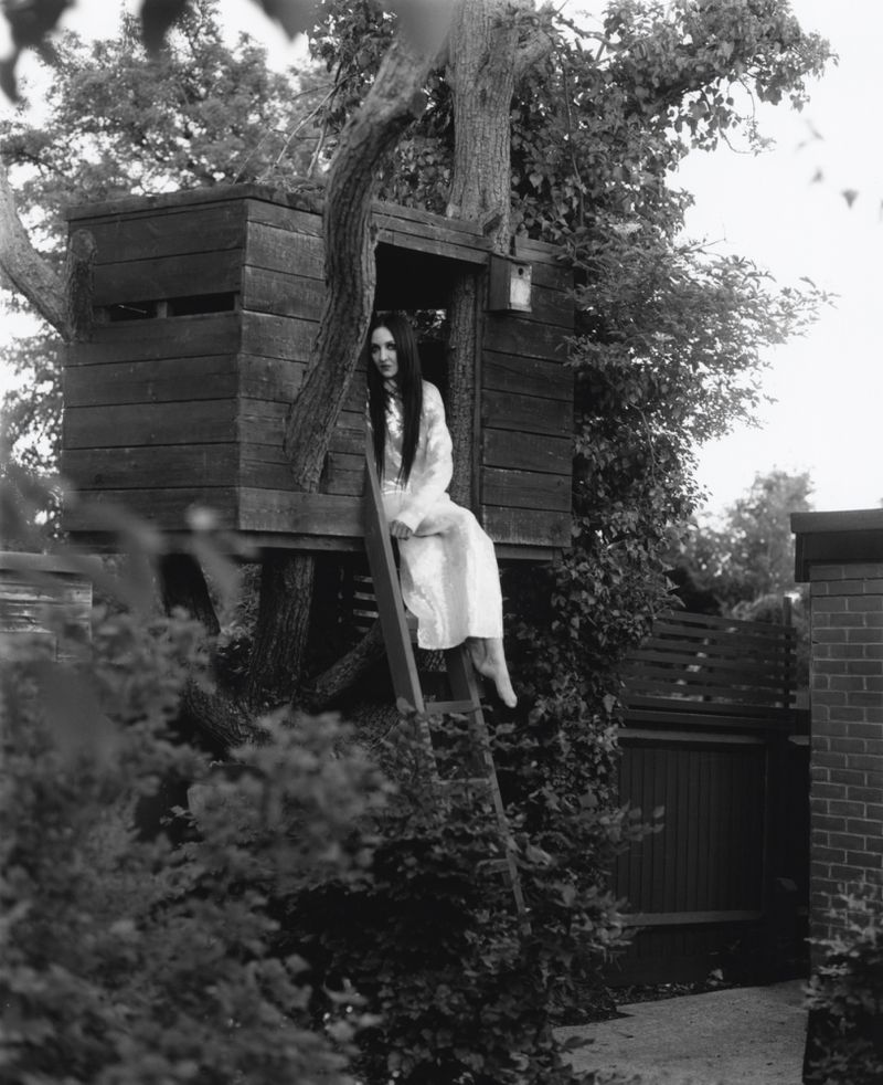 Tish Weinstock sitting in an elevated wooden treehouse in a garden wearing a long white dress in black and white.