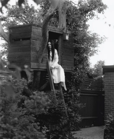 Tish Weinstock sitting in an elevated wooden treehouse in a garden wearing a long white dress in black and white.