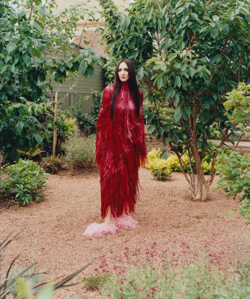 Tish Weinstock in a long red fringe gown and fluffy pink slippers standing on gravel in a garden surrounded by green foliage.