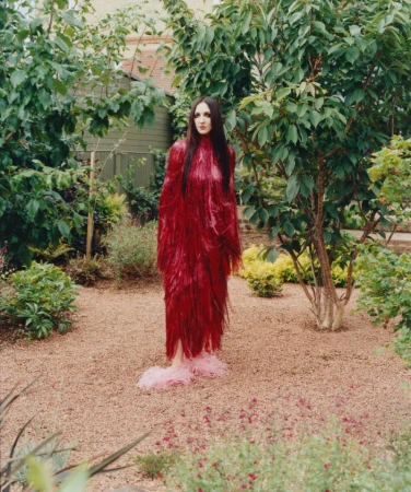 Tish Weinstock in a long red fringe gown and fluffy pink slippers standing on gravel in a garden surrounded by green foliage.