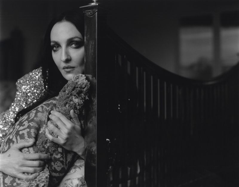Black and white portrait of Tish Weinstock peering past a dark wooden headboard while holding a patterned textile.