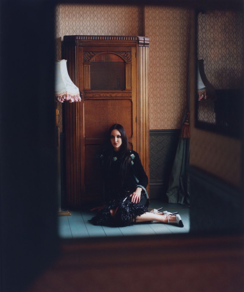 Tish Weinstock seated on the floor in a black embellished dress beside a wooden cabinet, seen through a dark framed doorway.