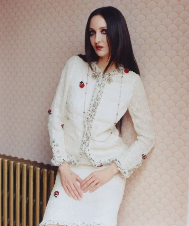 Tish Weinstock posing in an embellished cream tweed skirt suit against a patterned wallpaper wall beside a brass radiator.