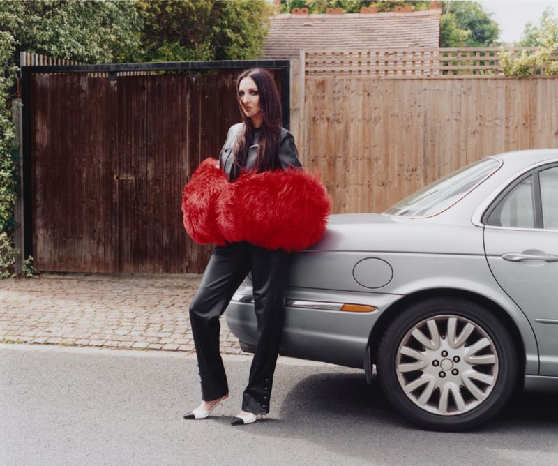 Tish Weinstock leaning on a silver car in black leather trousers and an oversized red faux fur stole on a quiet residential street.