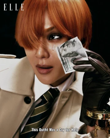 Close-up of person with short auburn hair wearing a trench coat and tie while holding a hundred dollar bill up to their face.