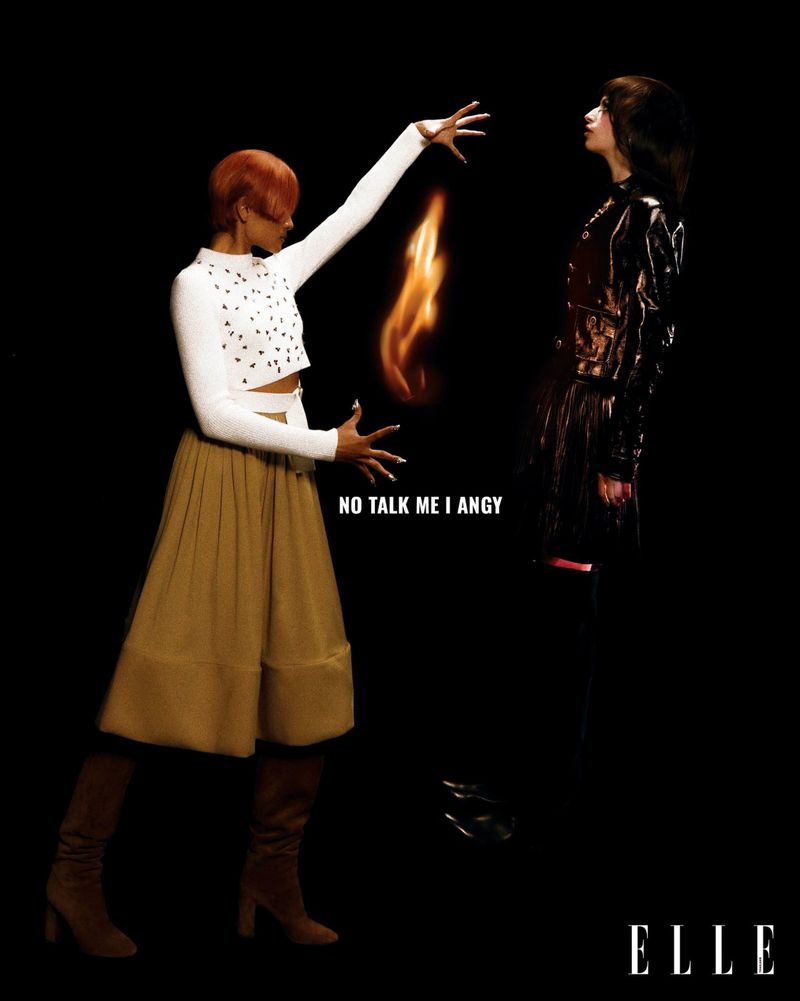Two women in a high-fashion, studio setting, one dressed in white and brown, appearing to conjure flames with the other in black