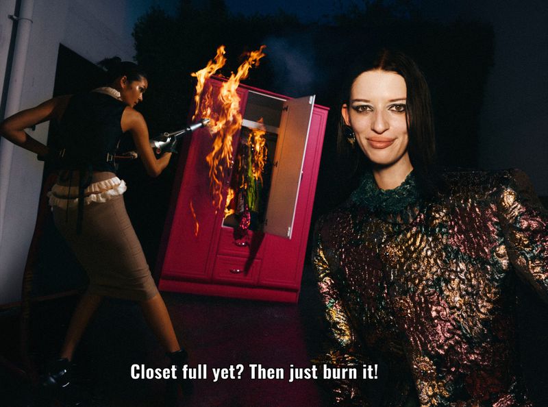 Two women pose beside a burning pink wardrobe in a darkly lit outdoor setting; one woman wields a flamethrower, creating a surreal fashion scene.