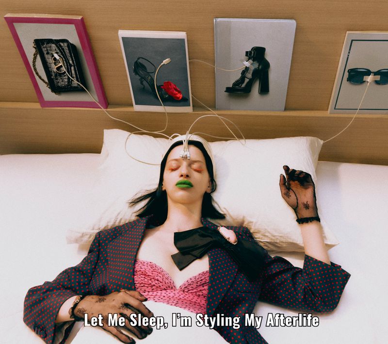 Model laying in bed, connected to wires, showcasing fashion accessories and makeup against a neutral-toned bedroom backdrop.