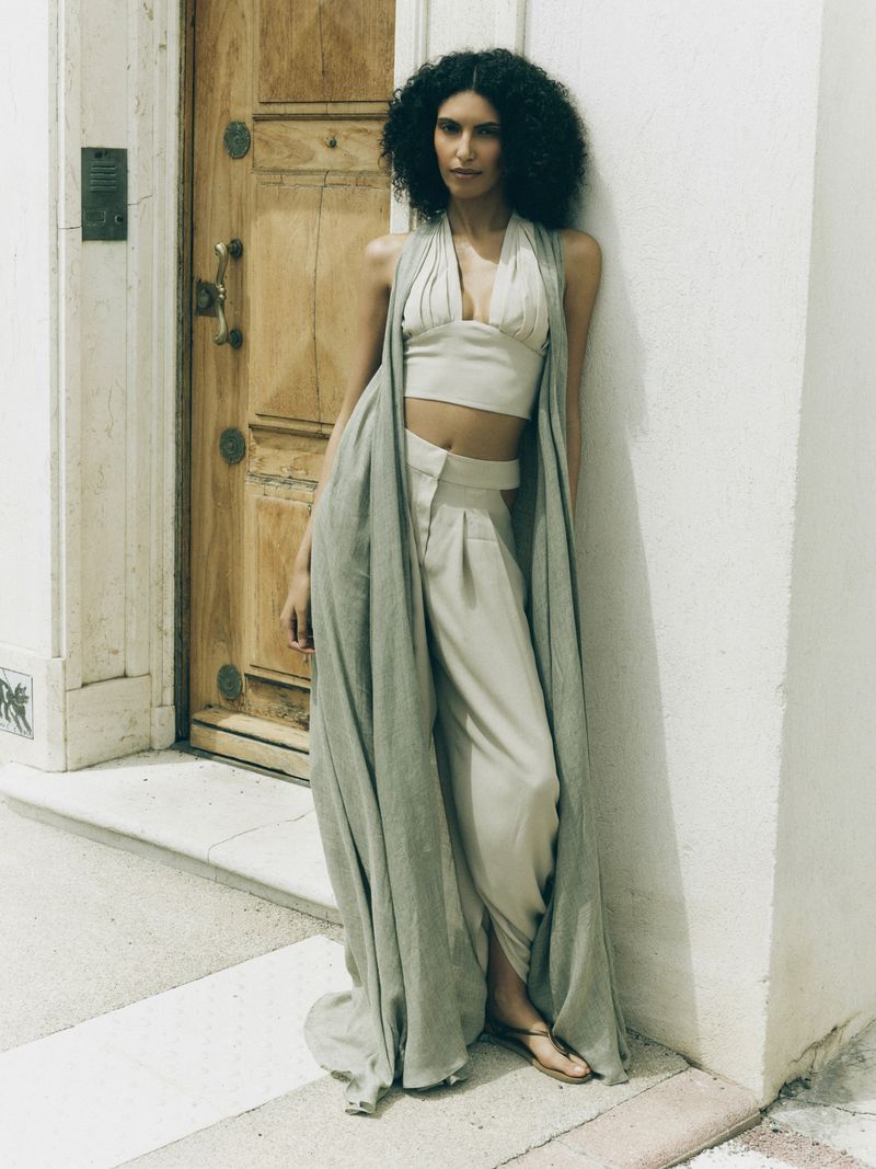 Model Sarah Ella wearing a draped sage vest and crop top leaning against a white exterior wall next to a wooden door.