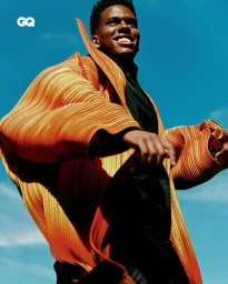 Estevão William wearing a textured pleated orange coat and black high-neck top smiling against a clear blue sky for GQ Brasil.