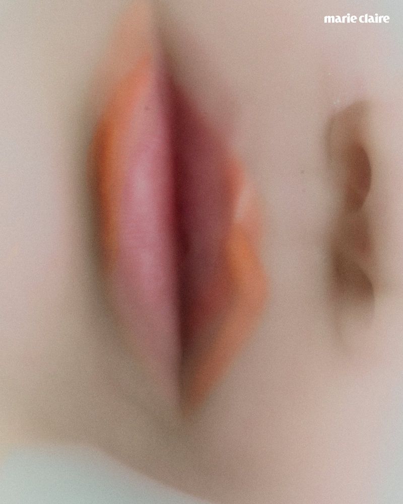 Macro abstract close-up of a doll face showing soft-focus orange painted lips and smooth pale skin with the Marie Claire logo.