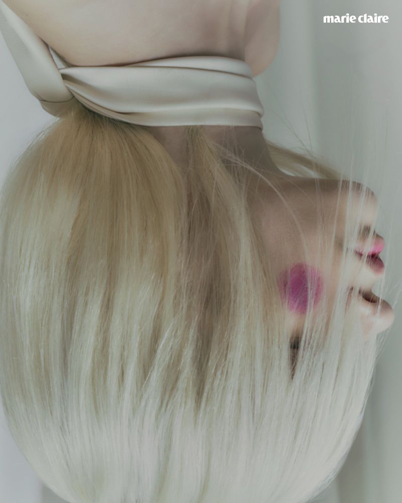 Close-up of a blonde model positioned upside down wearing a cream satin neck scarf with circular pink blush on her cheek.