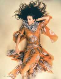Model Matilde de Nard lies down in an orange pleated dress with silver detailing and feather trim, her dark hair fanned out above her.