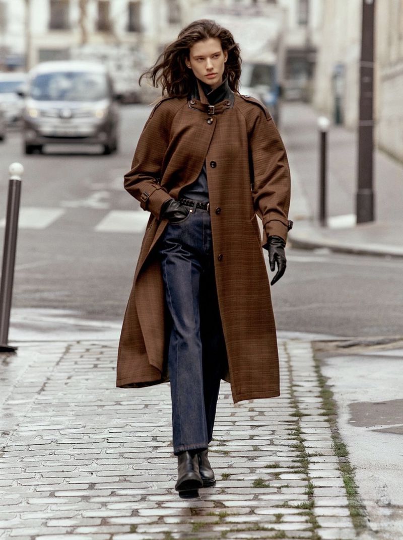 Model walking down a cobblestone street wearing an oversized brown coat, dark denim jeans, and black leather gloves.
